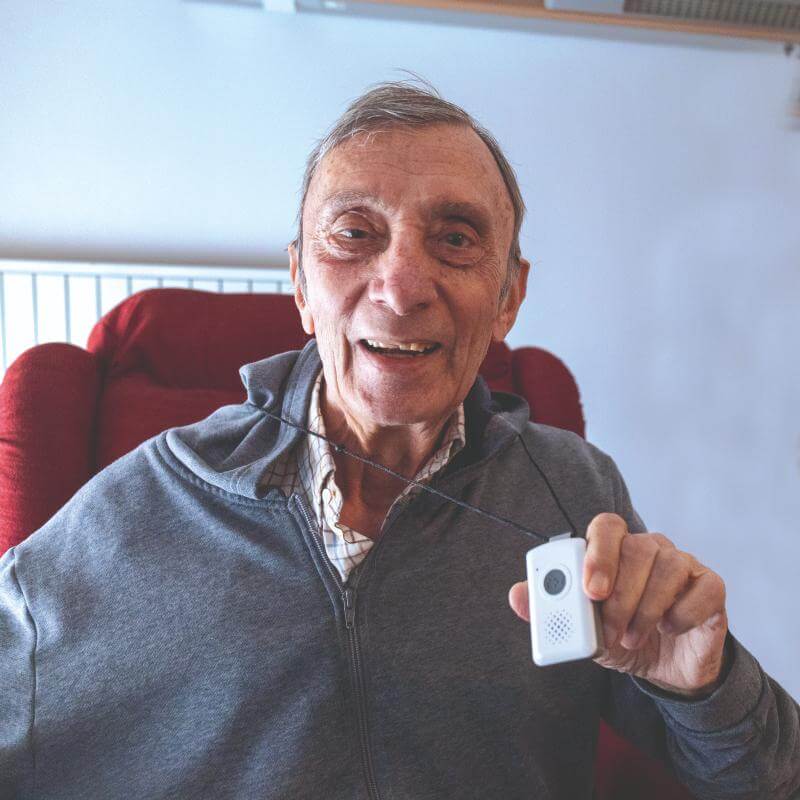 Luigi, cliente del telesoccorso, mostra con un grande sorriso il suo dispositivo alla telecamera.